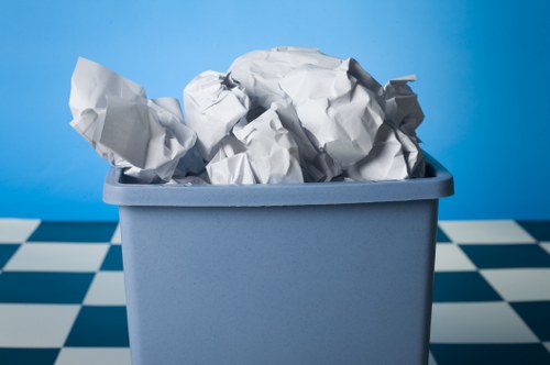 A light blue plastic bin filled with crumpled white paper and cardboard, placed on a checkered black and white floor with a plain blue wall in the background. The paper appears torn and wrinkled, with some pieces slightly leaning over the edge of the bin. The environment suggests an indoor space, possibly a storage area or part of a garden clearance setup, consistent with rubbish removal services in Brockley. The image focuses on the waste material being prepared or collected for disposal or collection, representing typical rubbish collection practices. Garden Clearance Brockley offers services that may include clearing such waste from residential or garden areas, and this image illustrates a bin filled with paper and cardboard waste ready for removal.