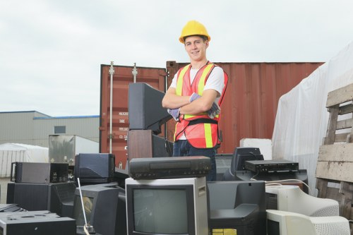 A smiling man wearing a yellow safety helmet and a high-visibility vest stands outdoors at a waste disposal site, surrounded by various discarded electronic devices including old computer monitors, desktops, and printers. The background features a large red shipping container and a stack of wooden pallets, with an overcast sky visible above. The scene conveys the process of rubbish removal, with the man appearing to oversee or coordinate the collection of electronic waste. The setting is characteristic of a commercial or industrial waste clearance environment, aligning with services offered by Garden Clearance Brockley in the Brockley area. The electronic items in the foreground display different textures and colours, primarily black, grey, and off-white, with some showing visible screens and casings. The overall atmosphere suggests a professional and organized rubbish clearance operation focused on electronic waste disposal.