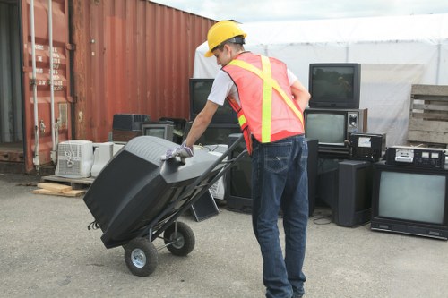 A person wearing a yellow safety helmet and a high-visibility orange vest with yellow reflective stripes is pushing a black wheeled waste container or bin, which is partially tilted on a paved outdoor surface. Behind them is a row of used electronic appliances, including televisions, monitors, and various small devices, placed against a background of red shipping containers and a wooden pallet. The scene likely takes place in a designated waste disposal or collection area, possibly related to garden or rubbish clearance services in Brockley, London, as suggested by the context of gardening and rubbish removal. The environment is well-lit, and the background shows a semi-industrial setting with an overcast sky, emphasizing the structured process of waste collection and recycling.