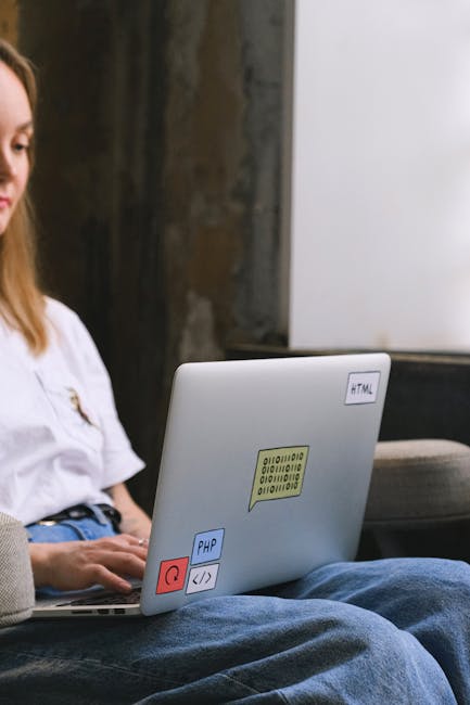 A person wearing a white shirt and blue jeans is seated on a beige fabric armchair, using a silver laptop that has several colorful stickers on its lid, including a software-related icon, a speech bub