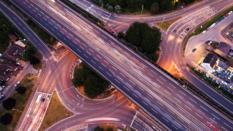An aerial view of a multi-level road interchange captured during dusk, showing blurred light trails from moving vehicles on the bridges and ramps, with some stationary cars parked in nearby residentia
